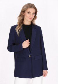 Námořnický modrý blazer s jednokřídlým zapínáním, dvěma předními kapsami a texturovanou látkou. Vyznačuje se štíhlým, upraveným střihem.