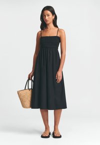 Vestido negro midi con tirantes finos, busto fruncido, falda fluida, combinado con un bolso de mimbre y sandalias planas. Textura suave.