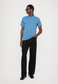 Modra kratka majica iz bombaža s kratkimi rokavi in krožnim ovratnikom, v kombinaciji z črnimi širokimi hlačami. Obutev je črna in sijajna, skupaj z okroglimi sončnimi očali.