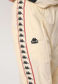 Bézs nadrág oldalsó csíkokkal, amelyeken ismétlődő fekete Kappa logók találhatók, valamint a Kappa logó a zseb közelében hímzett.