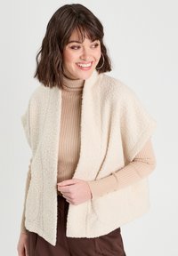 Gilet en fausse fourrure ivoire avec une ouverture devant, de forme surdimensionnée et avec des poches latérales, superposé sur un col roulé côtelé beige clair, ornée de boutons dorés.