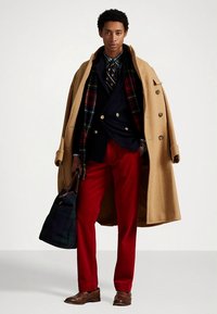 Brązowy wełniany płaszcz, granatowa dwurzędowa marynarka, czerwone spodnie, koszula w kratę, wzorzysty krawat, trzyma torbę w kratę. Brązowe skórzane buty.