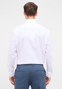 Camicia bianca a maniche lunghe con una texture liscia e colletto classico. Presenta polsini alla francese, abbinata a pantaloni blu e un design slim fit.
