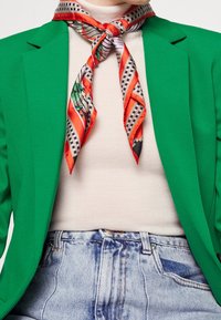 Blazer verde, dolcevita beige e pantaloni shorts in denim blu; sciarpa arancione e a fantasia legata al collo con quadretti e righe. Tessuto testurizzato.