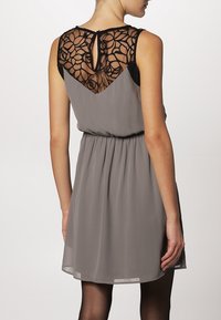 Graues Kleid mit taillierter Taille, fließendem Rock und einem schwarzen, gemusterten Spitzenrücken. Beinhaltet ein Schlüssel-Ausschnitt-Detail am Hals.