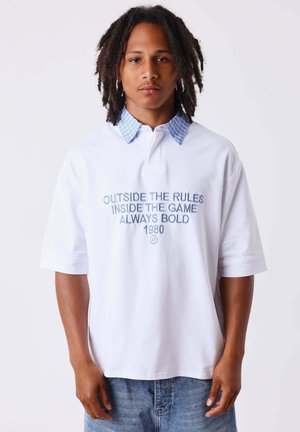 Jeune homme avec des dreadlocks portant un polo blanc avec un col rayé bleu et texte "Outside the rules inside the game always bold 1980."