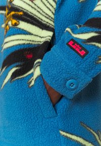 Manche de veste en polaire bleue avec motif graphique coloré, poignet boutonné et écusson logo rose et noir près du poignet.