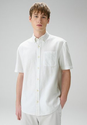 Noor mees, kes kannab valget lühikeste varrukatega nööbitavat särki rinnataskuga, seisab ühetaolise heledahalliga tausta ees.