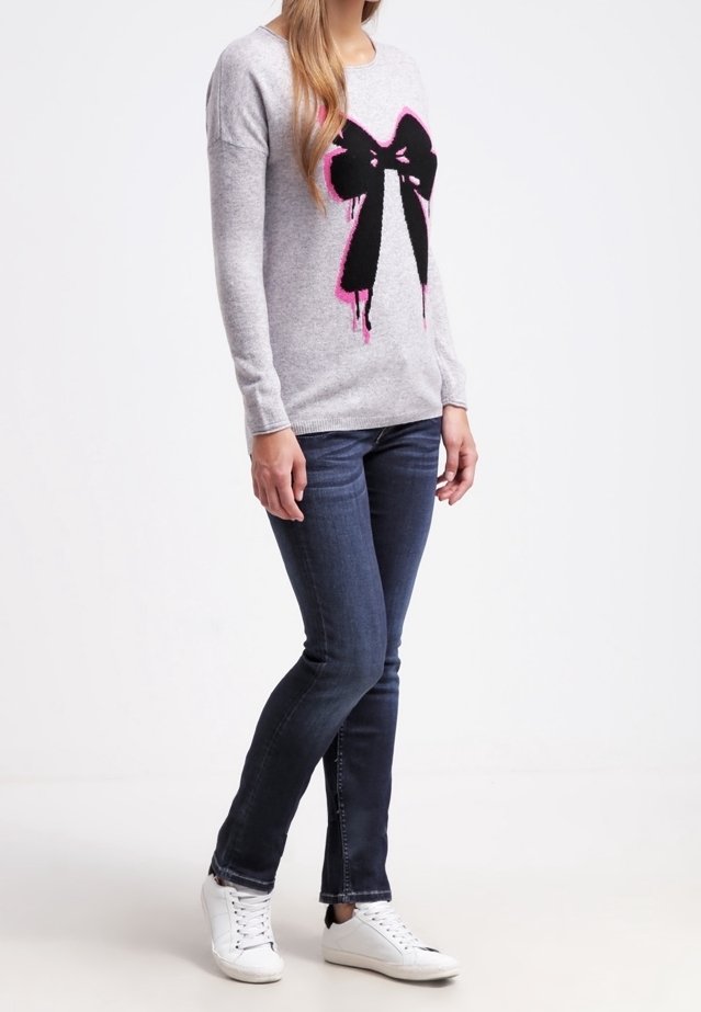 Suéter de punto gris con un gran gráfico de lazo negro y rosa, combinado con jeans oscuros y zapatillas blancas. Diseño sencillo con cuello redondo.