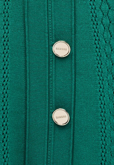 Tissu tricoté vert avec deux boutons en argent gravés du nom de la marque « SANDRO », présentant des motifs décoratifs en tricot le long des bords.
