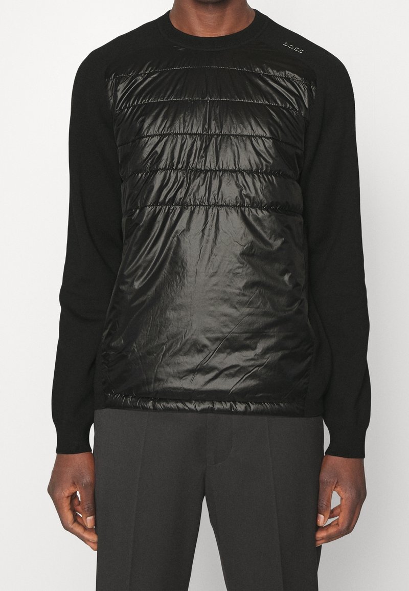 Czarny sweter z okrągłym dekoltem, z pikowaną, błyszczącą czarną panelą z przodu. Posiada ściągacze na mankietach i dyskretną markową metkę na ramieniu.