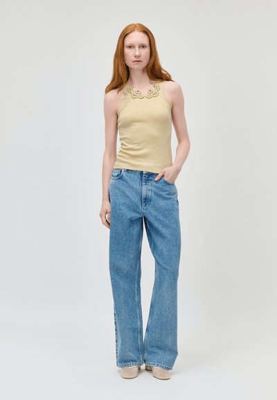 Femme aux longs cheveux roux portant un haut beige sans manches avec un détail en dentelle, un jean bleu ample et des chaussures beiges, debout avec une main dans la poche.