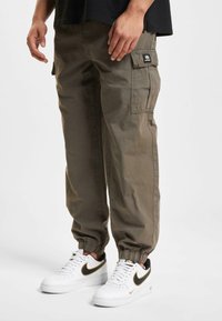 Olivegrønne cargo-bukser i bomuld med elastiske manchetter og sidelommer; båret med hvide sneakers med sorte og gyldne detaljer.