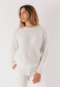 Donna con capelli biondi che indossa un maglione bianco a maglia e pantaloni bianchi, in piedi con una mano in tasca, che guarda a sinistra su uno sfondo neutro.