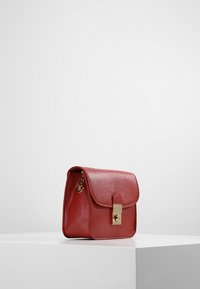 Červená kožená kabelka se strukturovaným tvarem, plochým klopovým víkem, zlatým zapínáním a bočním řetízkem. Hladký povrch se jemnou texturou.