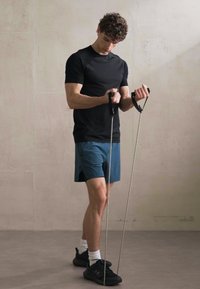 Camiseta deportiva negra de manga corta, pantalones cortos azules y zapatillas negras. Usuario sosteniendo bandas de resistencia, concentrado en su entrenamiento en un espacio minimalista.