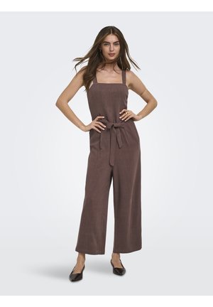 Femme debout, les mains sur les hanches, portant une combinaison marron sans manches à jambes larges avec une ceinture en tissu et des chaussures plates noires pointues sur fond clair.