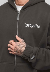 Dark gray zip-up hoodie with white "Dropsize" embroidery on the chest, ribbed cuffs, and a silver zipper. Textured fabric with a soft finish.