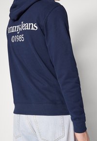 Námořnický modrý mikina s kapucí, která má klokankovou kapsu, žebrované manžety a pas. Na zadní straně je text "Tommy Jeans ©1985" v bílé barvě.