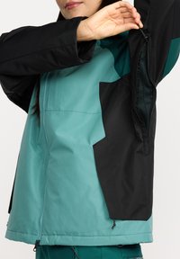 Veste teal et noire avec une poche zippée, texture lisse, col haut, et caractéristiques ajustables à l'ourlet et aux poignets.