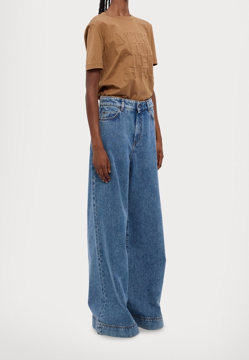 Personne portant une chemise marron à manches courtes et un jean bleu taille haute à jambes larges, debout devant un fond uni.