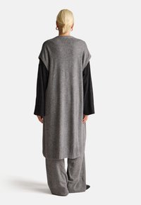 Dlouhý šedý pletený kardigan s volným střihem, s žebrovaným lemem, spárovaný s odpovídajícími širokými kalhotami a černými dlouhými rukávy pod ním.