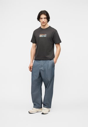 Mladý muž s tmavými vlasmi, ktorý má na sebe čierne tričko s potlačou One Piece, voľné modré rifle a svetlo béžové tenisky, stojaci s jednou rukou v vrecku.