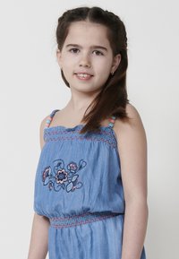 Džinsinis suknelė su raukiniuotu liemeniu, rožinių ir mėlynų siuvinėtų gėlių raštu, ir karoliukais papuoštais petnešomis. Audinys turi švelnų tekstūrą.