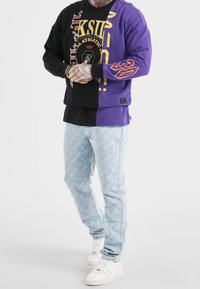 En person iklädd en svart och lila delad sweatshirt med grafisk text, ljusblå mönstrade jeans och vita sneakers, står med händerna ihopknäppta.