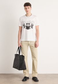 Hvit bomulls T-skjorte med svart grafisk trykk av løvehoder, paret med beige bukser og en svart tote-bag preget med "HUGO."
