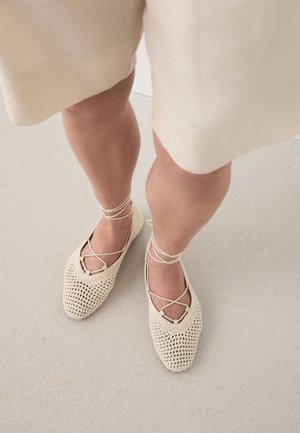 Benen met beige knielange shorts en crèmekleurige gevlochten veterschoenen op een lichtgrijze vloer.