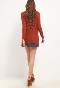 Maglione lungo color ruggine con un tessuto lavorato e un intricato design con pizzo sul retro. Abbinato a una gonna di jeans e tacchi alti marroni.