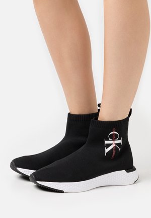 Stivaletti alla caviglia neri in maglia con suola in gomma bianca. Presentano un logo in rosso e bianco. Design alto con polsino a coste per una vestibilità aderente.