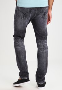 Jeans de mezclilla gris oscuro con textura deslavada, corte slim y bolsillos traseros distintivos con costuras. Combinados con zapatos negros sin cordones.