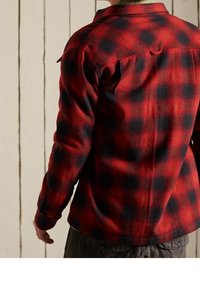 Camisa de franela con cuadros rojos y negros, mangas largas, costuras visibles, detalle de botón en el cuello, tela texturizada, dobladillo curvado.