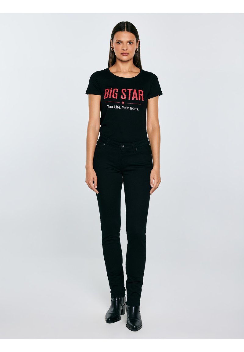 Nő áll fehér háttér előtt, fekete "BIG STAR Your Life. Your Jeans." feliratú pólót, fekete farmert és fekete bokacsizmát visel.