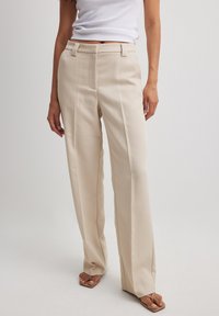 Beige tailored trousers with a straight-leg silhouette, structured waistband, and subtle texture. Worn with a fitted white top.