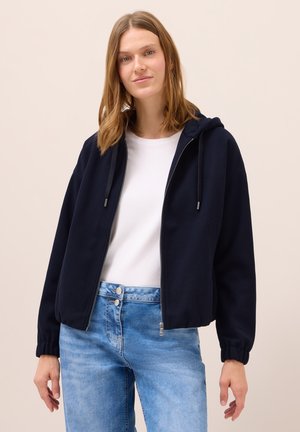 Veste à capuche zippée navy avec une coupe décontractée, dotée de poignets côtelés et d'une couleur unie, associée à un t-shirt blanc et un jean bleu clair.