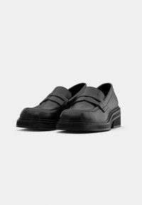 Paire de mocassins noirs en cuir à enfiler avec bout rond et semelle épaisse sur un fond uni gris clair.
