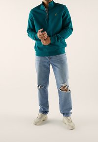 Sininen vetoketjullinen neulepaita resoreilla varustetuilla hihoilla, yhdistetty vaaleansinisiin repeytyneisiin farkkuihin ja beigen värisiin lenkkareihin. Neulepaidassa on pieni keltainen logo.