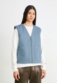 Gilet en coton bleu avec fermeture éclair sur le devant, côtés ouverts, deux poches avant et un accent avec un logo. Porté sur une chemise à manches longues blanche et un pantalon marron.