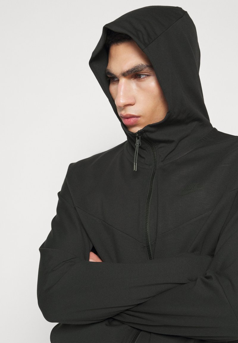 Felpa nera con zip, colletto alto, cappuccio e design a chevron. Realizzata in tessuto liscio e leggero. Presenta un logo sottile sul petto.
