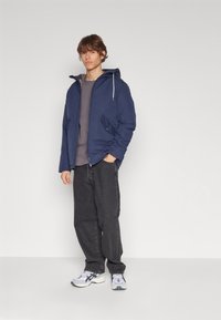 Veste à capuche bleu marine avec poches avant, associée à un sweat-shirt gris et un jean noir. Les chaussures sont de style athlétique gris et blanc.