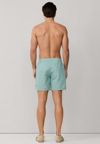 Modelo masculino de pie de espaldas, vistiendo pantalones cortos de baño azul claro y zapatos beige sin cordones, sobre un fondo liso gris claro.
