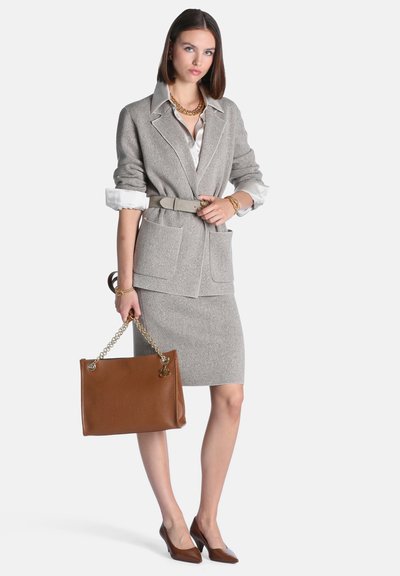 Femme en blazer gris et jupe avec chemise blanche, tenant un sac à main marron avec des anses en chaîne, portant des bijoux en or et des talons marron.