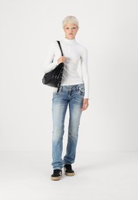 White long-sleeve top, light blue jeans, and black sneakers. Model carries a black bag with silver accents. Simple, fitted design.