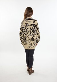 Maglione lavorato a maglia beige con motivo paisley nero, design oversize, scollatura a costine e spalle basse, abbinato a jeans skinny neri e stivali marroni.