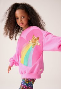 Roza majica z velikim grafičnim motivom srca, zlatno zvezdo iz kamenčkov in mavričnim vzorcem. Model nosi barvite legice z vzorcem zvezd.