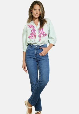Light green striped button-up shirt with pink floral embroidery, paired with blue jeans and beige shoes. Model stands with hands on hips.