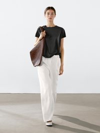 Sort bomulds t-shirt, hvide ribbede bukser med vid ben, brun flettet tote bag og hvide sko; kvinde i en minimalistisk studioindstilling.